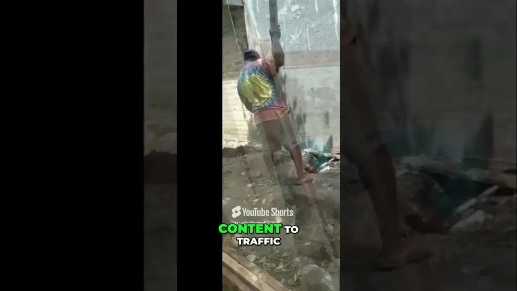 Man in a tie-dye shirt climbs a rope attached to a wall at a rough, unfinished construction site with debris around him.