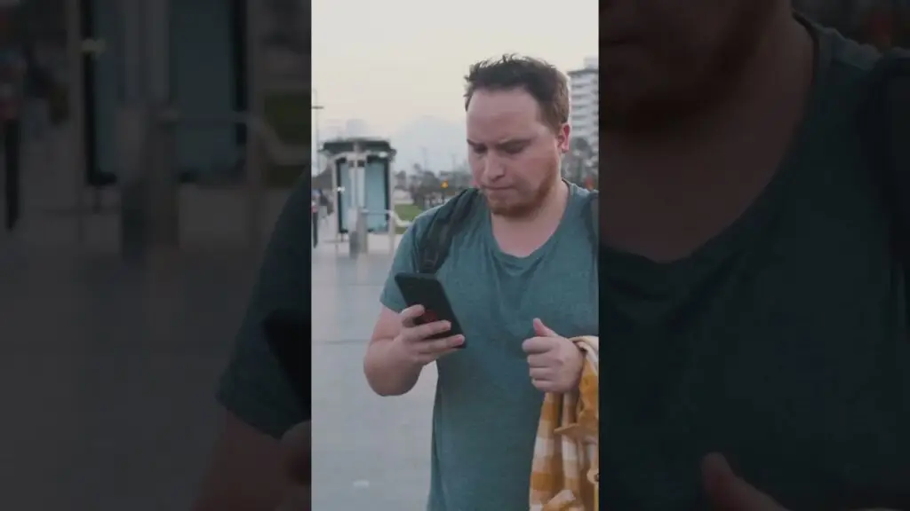Man with a backpack wearing a teal shirt looks down at his smartphone outdoors in an urban area,”