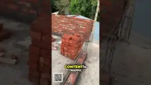 Stack of red bricks at a construction site beside a partially built brick wall with vertical rebar and surrounding greenery in the background.