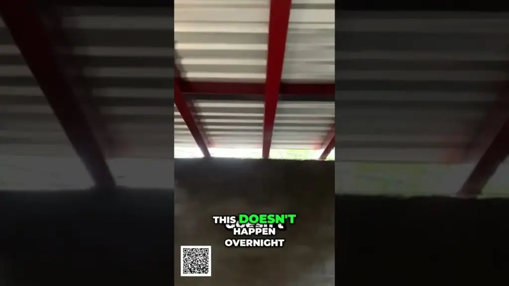 Video frame showing a dark industrial interior with red metal beams; white-and-green text reads 'THIS DOESN'T HAPPEN OVERNIGHT' and a QR code is at the bottom left.