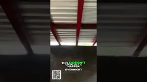 Video frame showing a dark industrial interior with red metal beams; white-and-green text reads 'THIS DOESN'T HAPPEN OVERNIGHT' and a QR code is at the bottom left.
