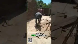 Construction site: worker in a gray shirt cutting a wooden plank on the ground with a power saw; scattered boards around and brick walls in background.