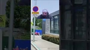 Video still of a man wearing a star-pattern bandana sitting on a city sidewalk near a modern building and a no-parking sign in the background.
