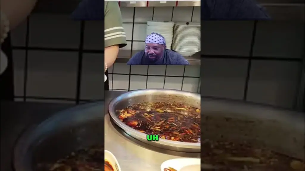 Man wearing a stars-pattern bandana looks down at a large simmering pot of spicy broth in a stainless steel kitchen. Caption 'UH' in green text appears over the pot.