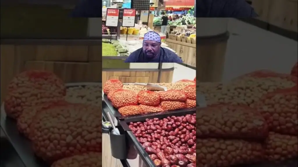 Man in a grocery store looking at a produce display with piles of red dates and orange fruit in the foreground/counter behind him.