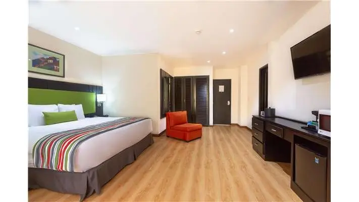 Hotel room featuring a king bed with a green headboard and striped runner, a red armchair, wood flooring, and a desk with a mounted TV and mini-fridge in the background.
