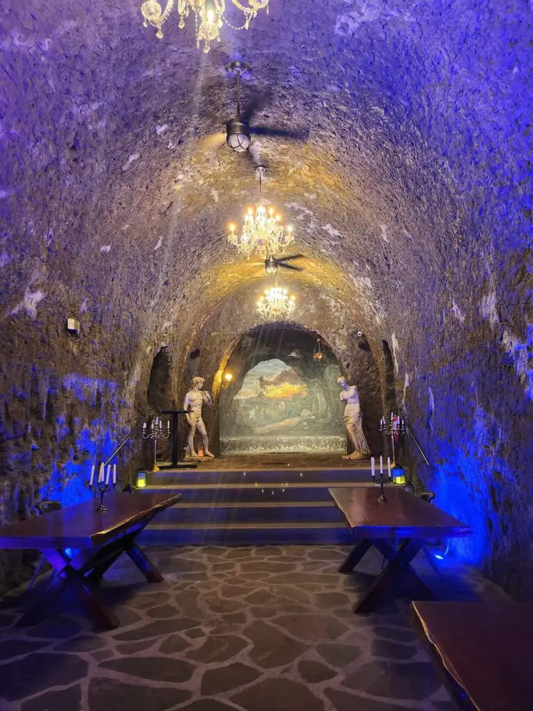 Underground stone hall with arched ceiling, two statues, and chandeliers; stone floor with wooden tables and candle holders illuminated by blue and warm lights.