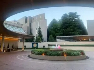 twg-new-sanno-hotel - Travel With Glen - Travel With Glen Circular plaza with a central lit fountain surrounded by flowers, under a curved architectural overhang and modern buildings in the background.