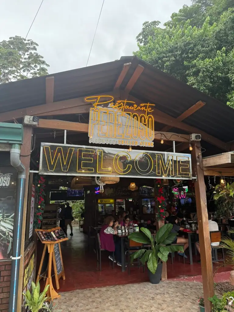 twg-restaurant-el-perezoso - Travel With Glen Open-front restaurant Perezoso with a neon 'PEREZOSO' sign and a large 'WELCOME' sign; customers dine at tables under a wooden roof.
