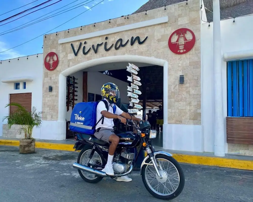 Delivery rider on a Suzuki motorcycle with a large blue insulated bag, parked in front of a storefront labeled 'Vivian'.
