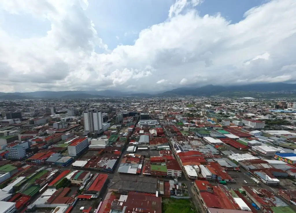 Aerial view of a densely built city with colorful red, green, and blue roofs, mountains on the horizon and a cloudy sky above.
