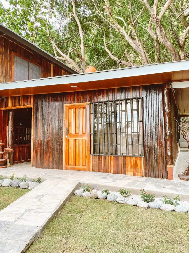 Wooden cottage-style house with an orange front door and a barred window, set beside a stone walkway and small garden. Behind it are large tree branches and green foliage.